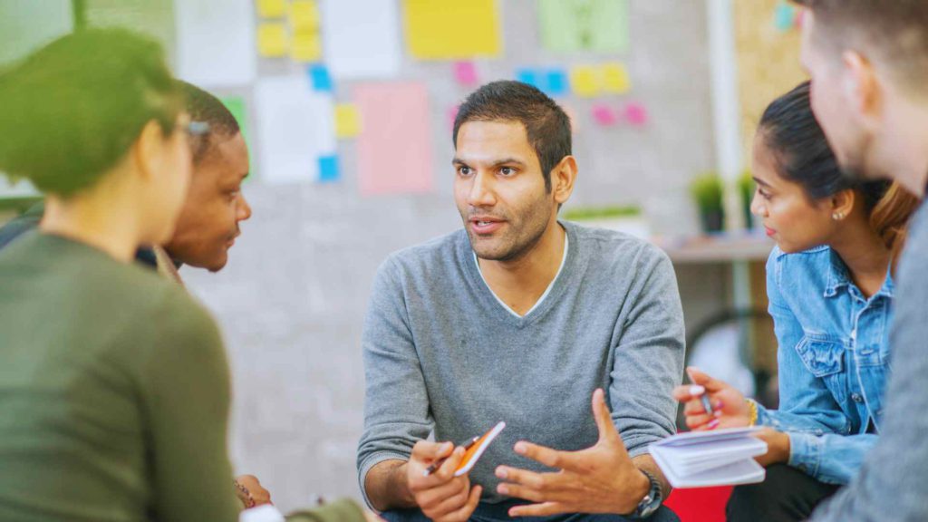 Team of people at work around a table discussing innovation, with sticky notes on the wall behind them